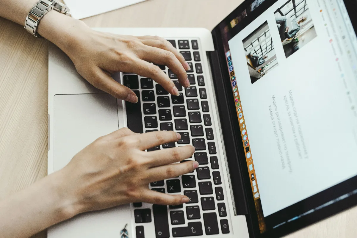 Overhead view of two hands typing on a laptop keyboard with an article and photo thumbnails on the screen