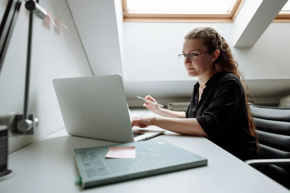 A woman in a black shirt working at a laptop beside a bright window, a notebook and pen on the white desk